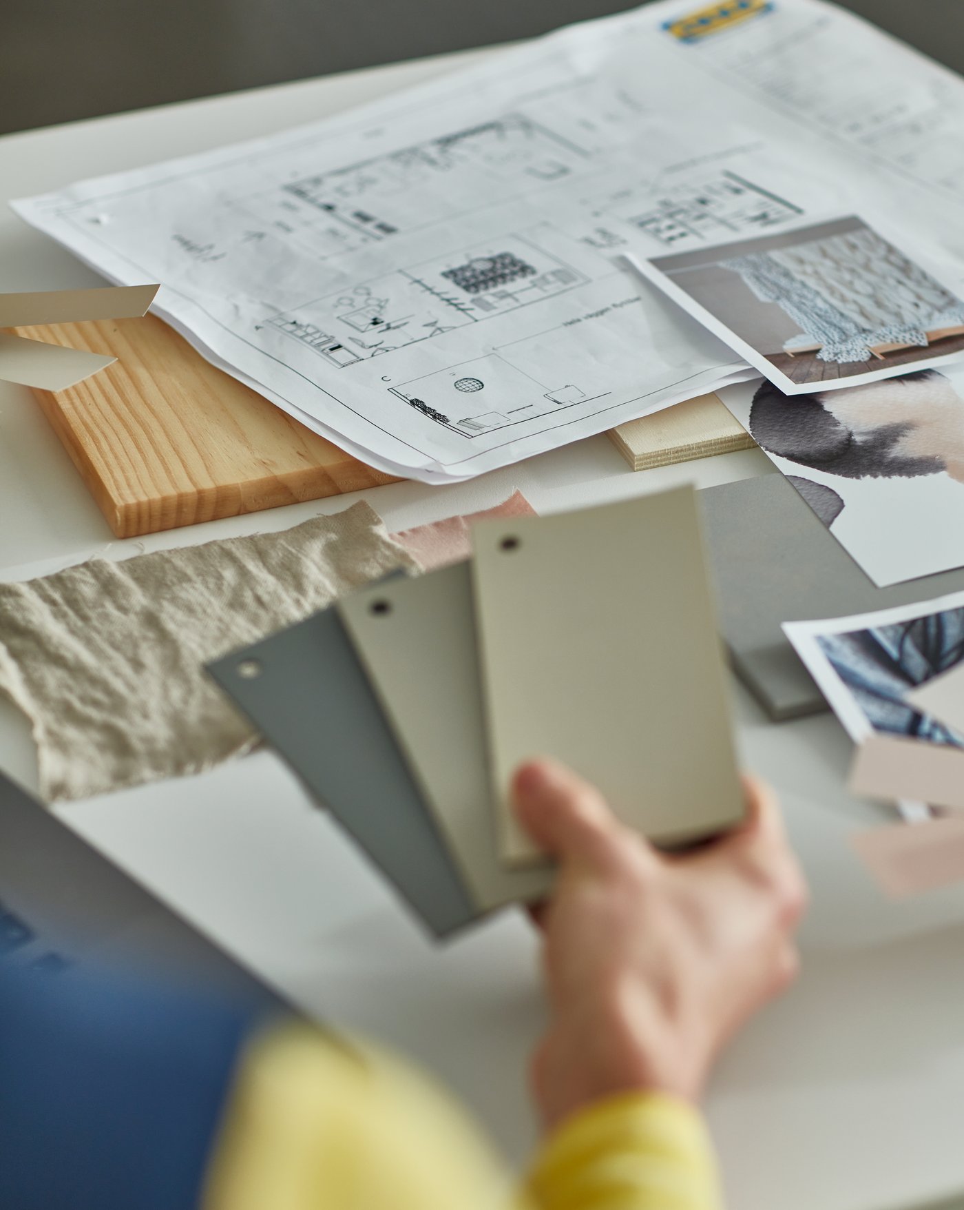 A hand of an IKEA interior designer holding some colour samples on top of a table full of design drawings.