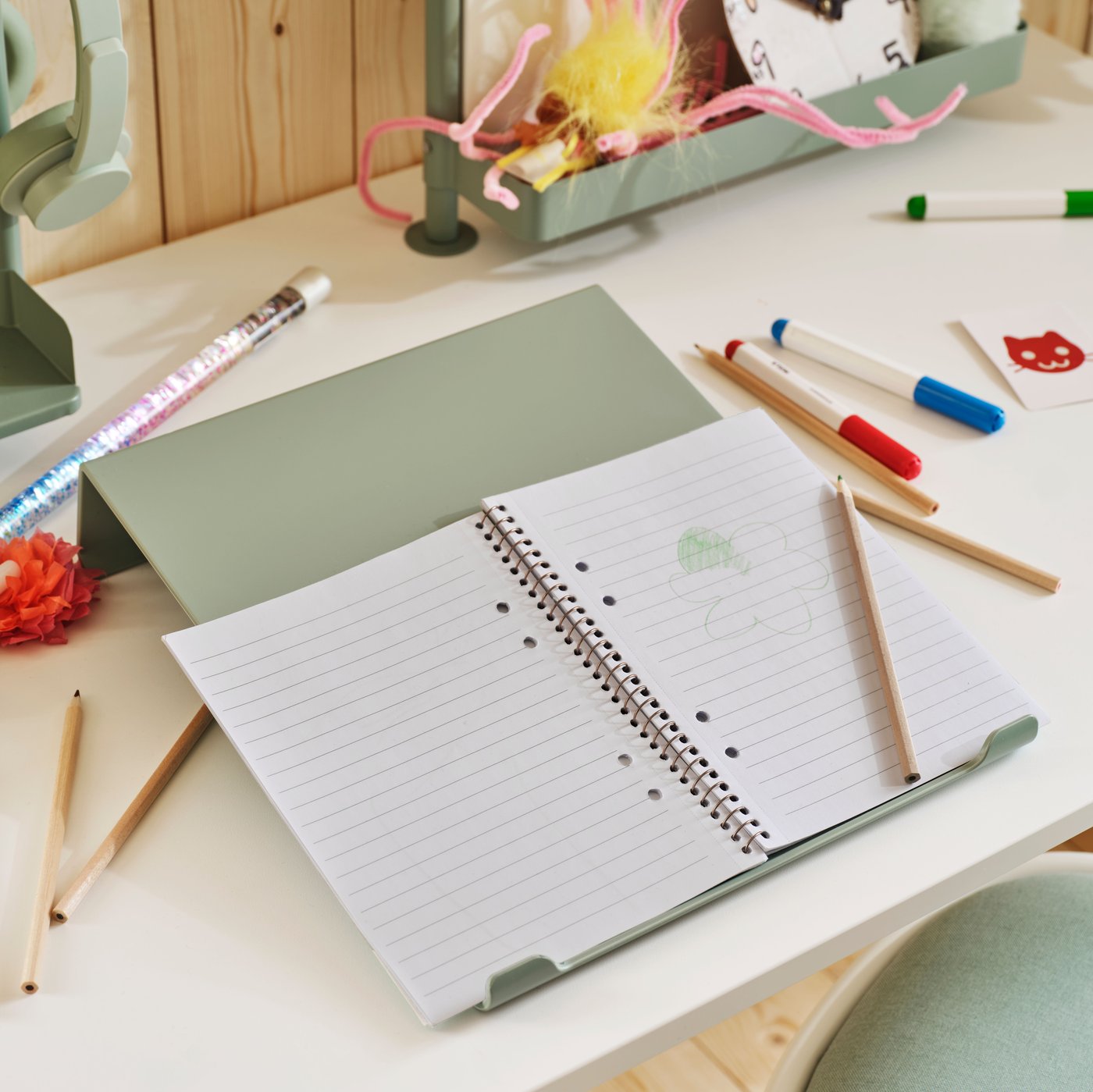 Pupitre d’écriture RELATERA en gris-vert clair sur un bureau assis/debout RELATERA blanc. Un cahier ouvert est posé sur le pupitre d’écriture, près de crayons épars.