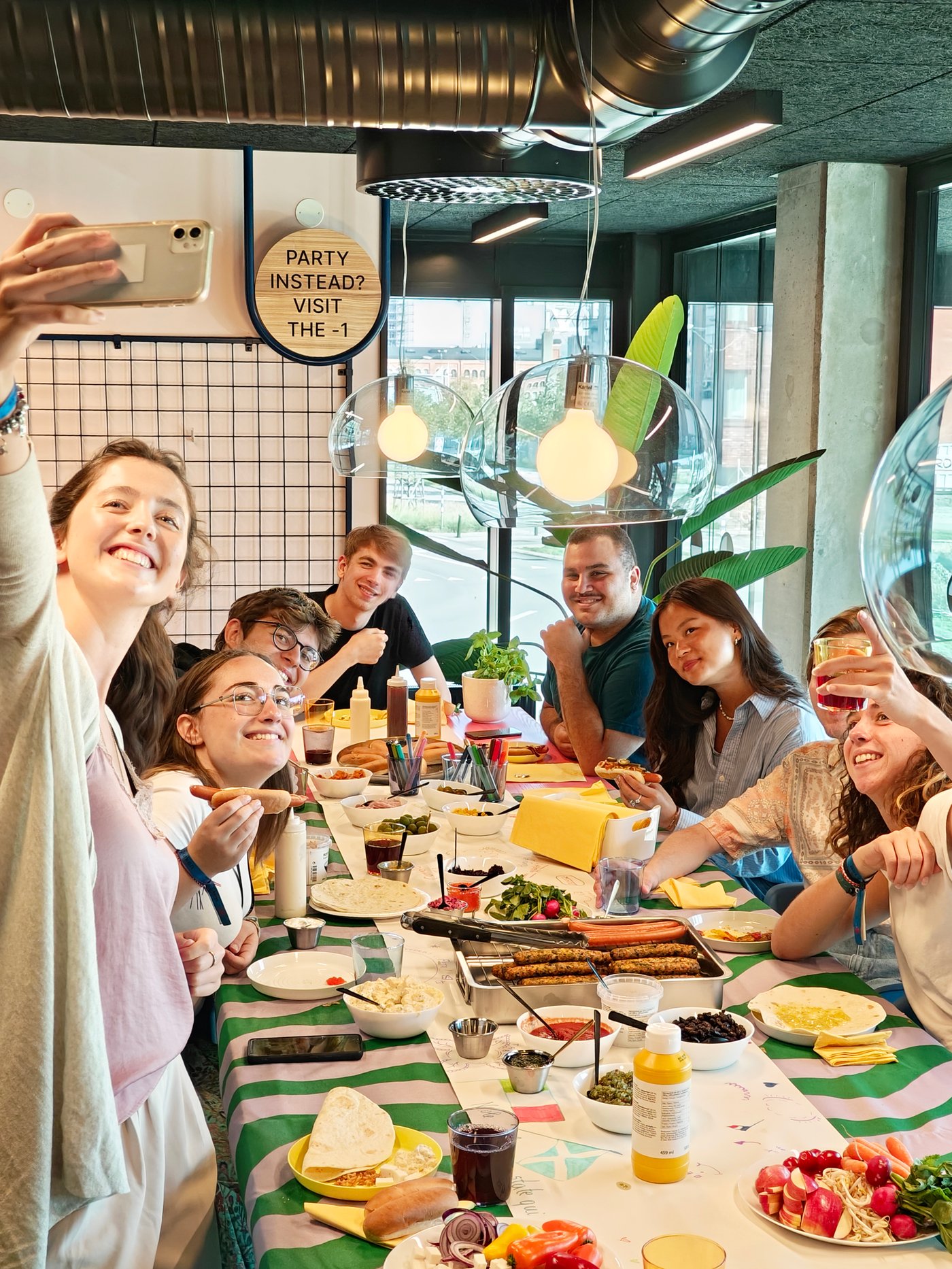 A group of young people gathered around a dining table eating hot-dogs.