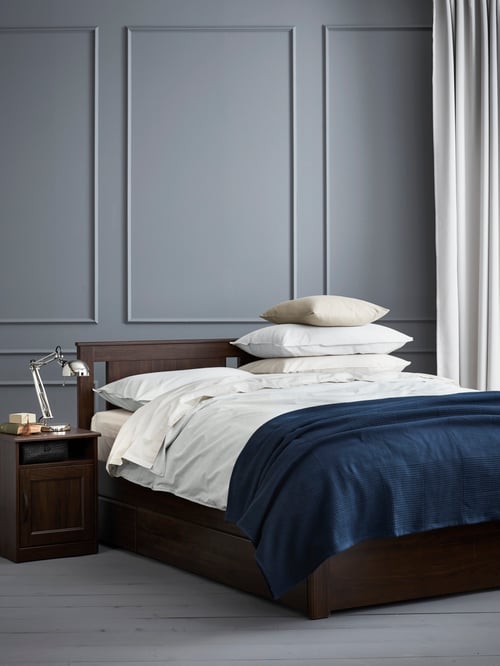 Bedroom with grey walls, brown bed frame with four storage boxes, white bed linens, blue bedspread, and brown bedside table.