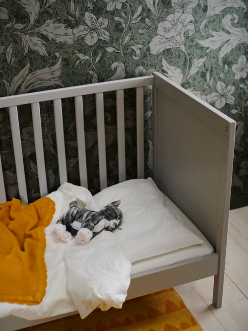 A LILLEPLUTT soft toy cat lies near a SOLGUL blanket in a SUNDVIK cot with LENAST bed linen. The cot has one side removed.