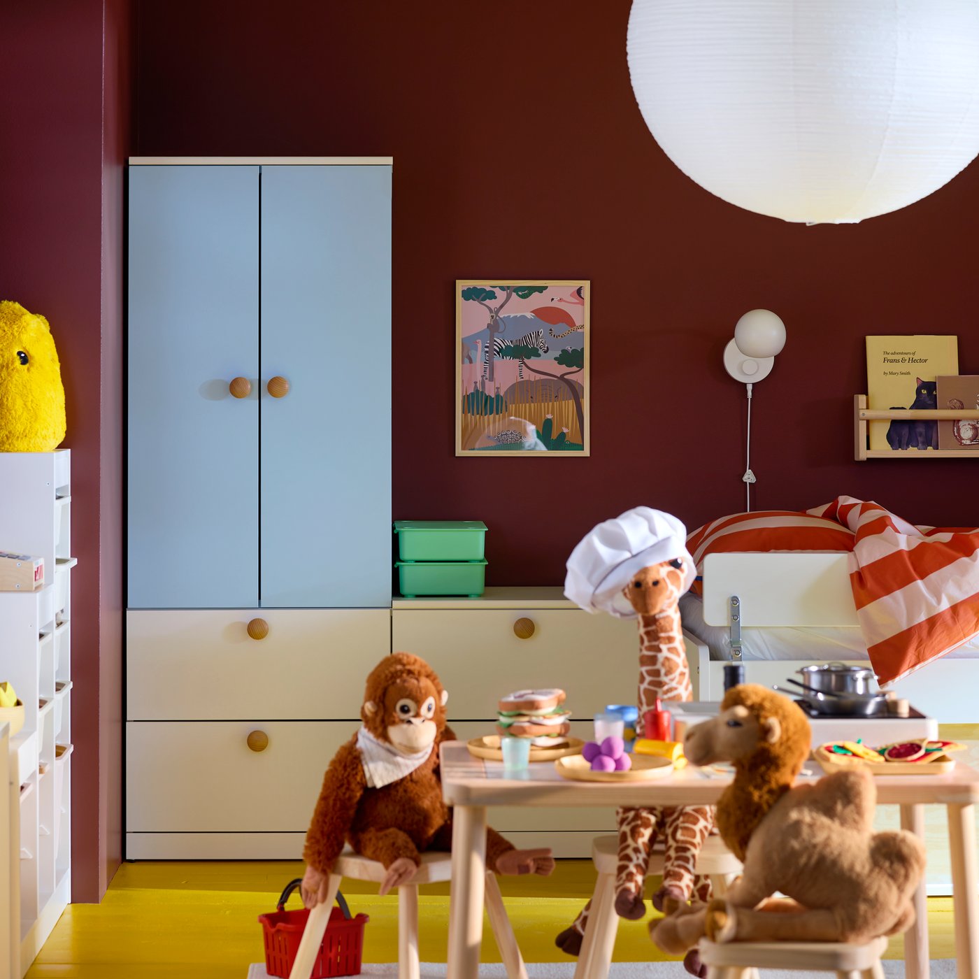 Une table FLISAT en bois clair dans une chambre d'enfant avec des peluches en train de cuisiner, une armoire bleue LASTARE et un mur brun-rouge.