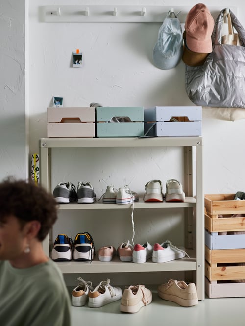 A hallway with three wooden boxes painted in different colours on top of a beige KOLBJÖRN shelving unit with shoes inside.