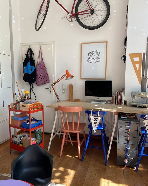 Blick auf den Schreibtisch mit Computer und Kreativbereich. Ein Fahrrad hängt an der Wand über dem Schreibtisch.