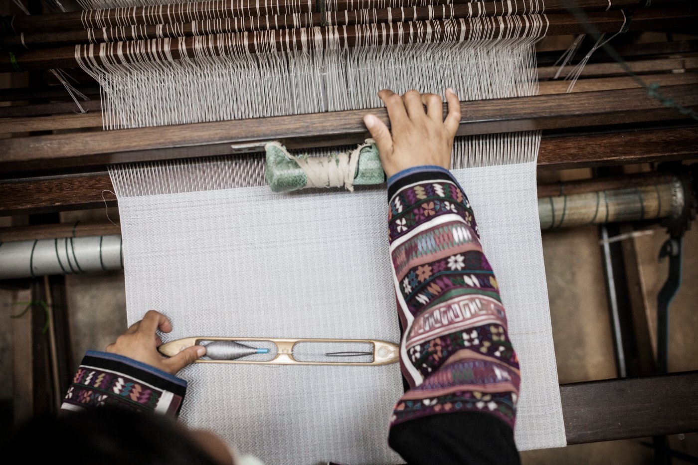 En person väver tyg på en vävstol. Personens händer är synliga och håller en skyttel medan de styr trådarna. Vävstolen har flera trådar spända över den, och personen bär kläder med intrikata mönster på ärmarna.