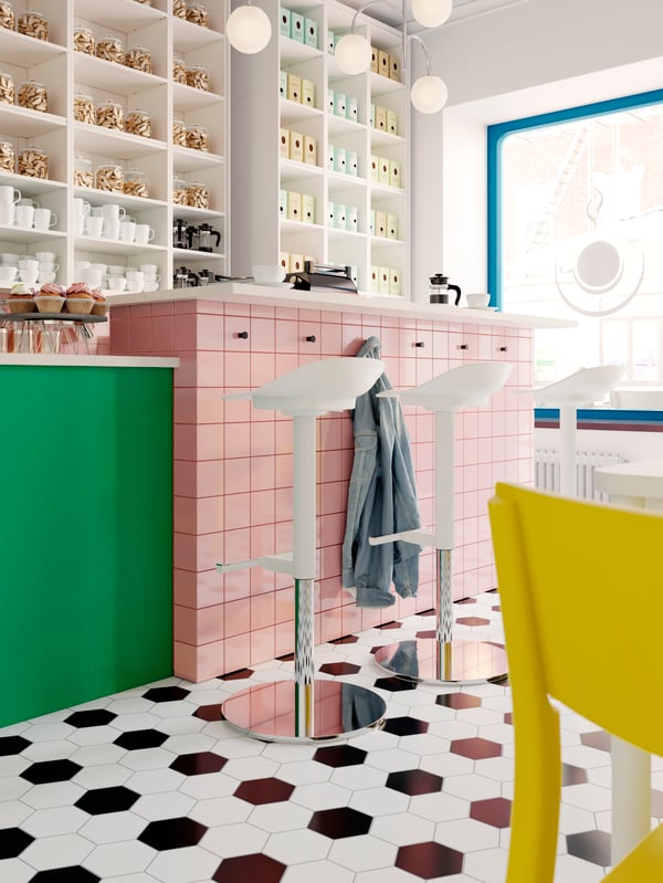 A light coffee-bar interior with floor-to-ceiling white shelves and white JANINGE bar stools by a counter.