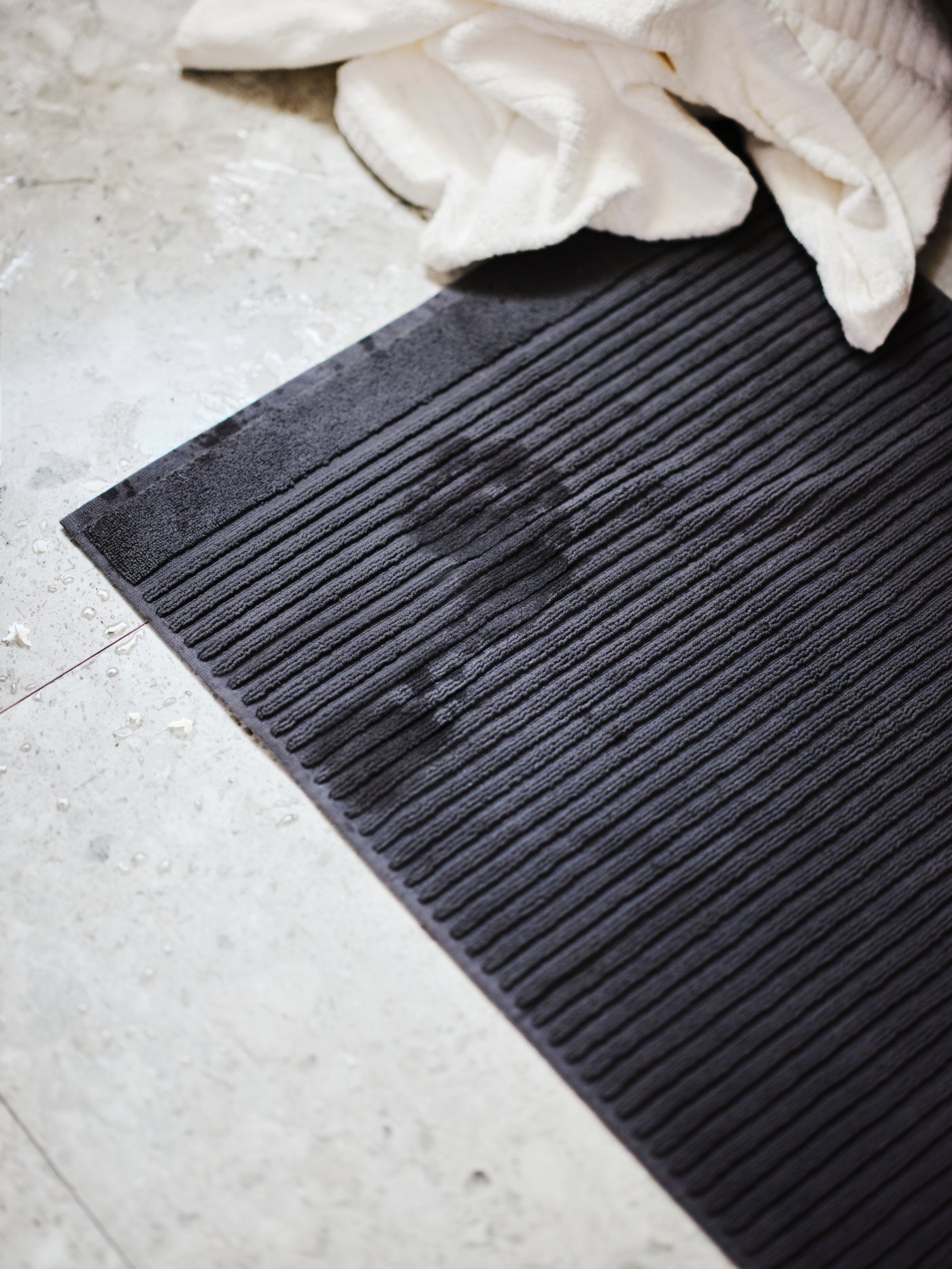 Tamna rebrasta kupaonska prostirka na svijetlom podu, s vidljivim mokrim otiscima stopala i bijelim ručnikom položenim uz rub.