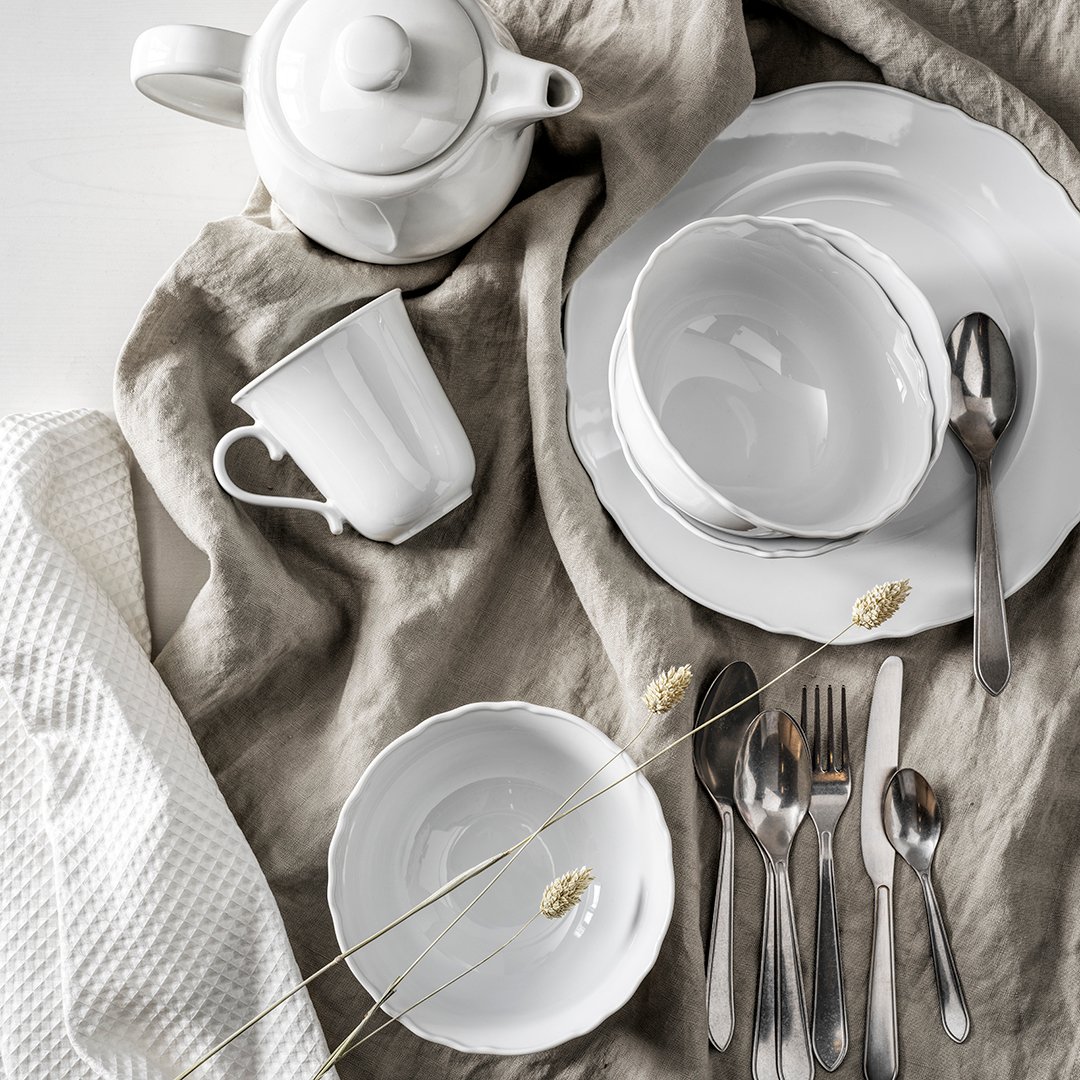 Et flot dækket bord i hvide og beige farver med tallerkner, bestik, glas og en stofserviet.
