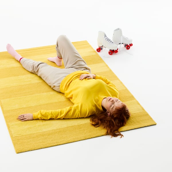 A person is lying comfortably on the floor, on top of a yellow LANDSVÄG rug. Next to the rug, a pair of roller-skates.