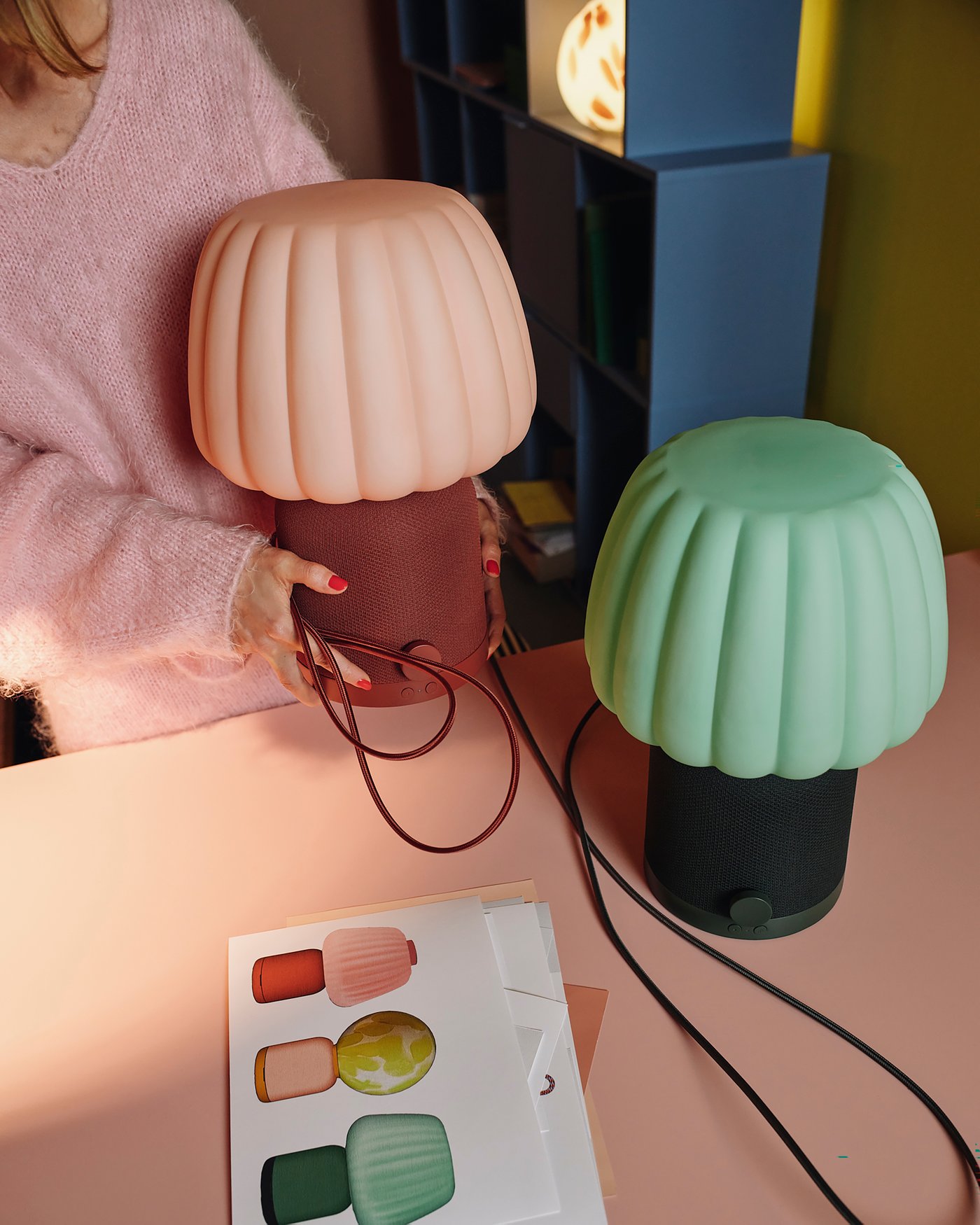 Two KULGLASS table lamps/speakers with pleated shades, one pink and one green, stand on a pink table. A person is holding the pink lamp, and a brochure with lamp pictures is next to it.
