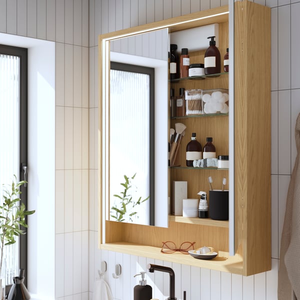 An oak effect FAXÄLVEN mirror cabinet with built-in lighting and two mirror doors has a door open, showing toiletries inside.