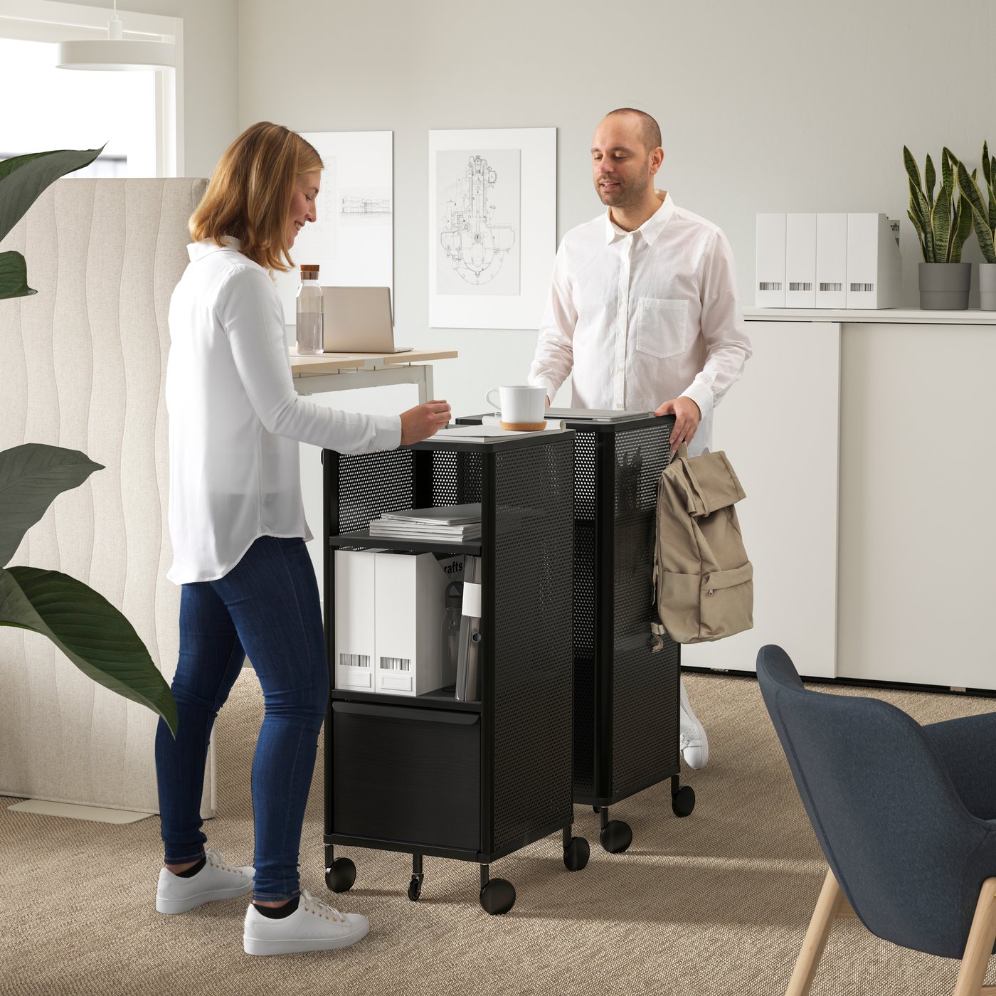 Two people gather at a mesh/black BEKANT storage unit on castors.