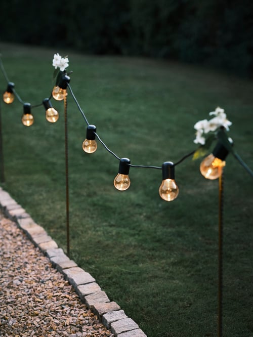 Focused shot of a lit SVARTRÅ lighting chain hanging from thin poles on top of which are white flowers. 