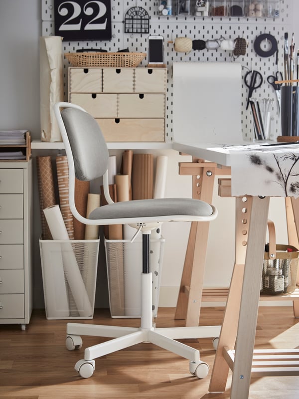 Una silla giratoria en blanco/Vissle gris claro, un escritorio blanco con caballetes de madera y dos cestas blancas con rollos de papel de estraza.