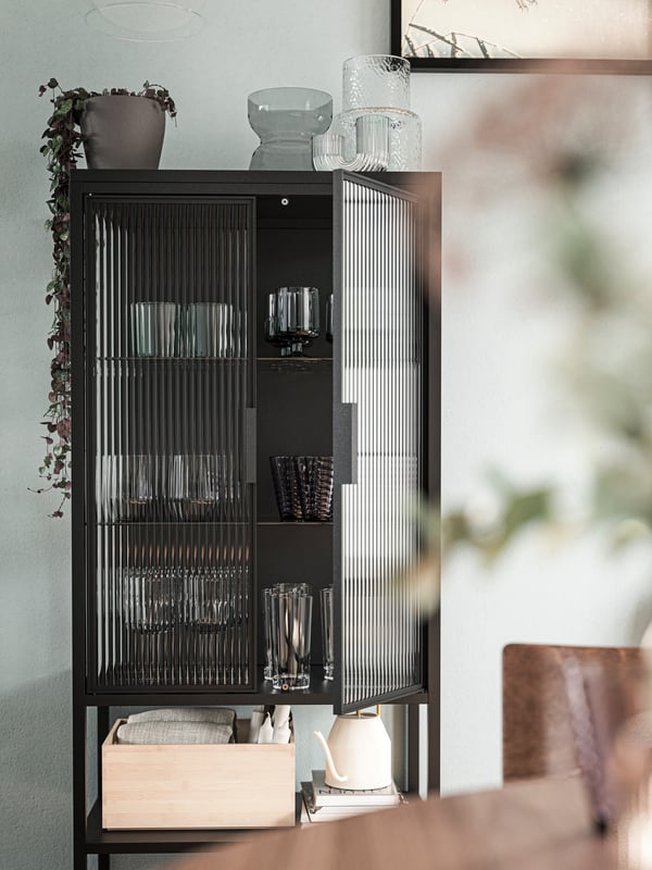 A MOSSJÖN glass-door cabinet with 2 doors holding glassware, an UPPDATERA storage box and a VATTENKRASSE watering can.