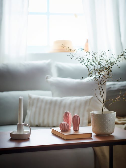 Living room with a white candlestick, a pink decoration set, and a potted plant on a table. A sofa is in the background.