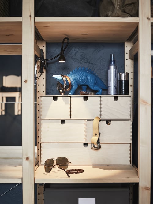 Close up on bedroom corner with shelf combination holding a mini wooden chest of drawers, toys, accessories, clamp spotlight.