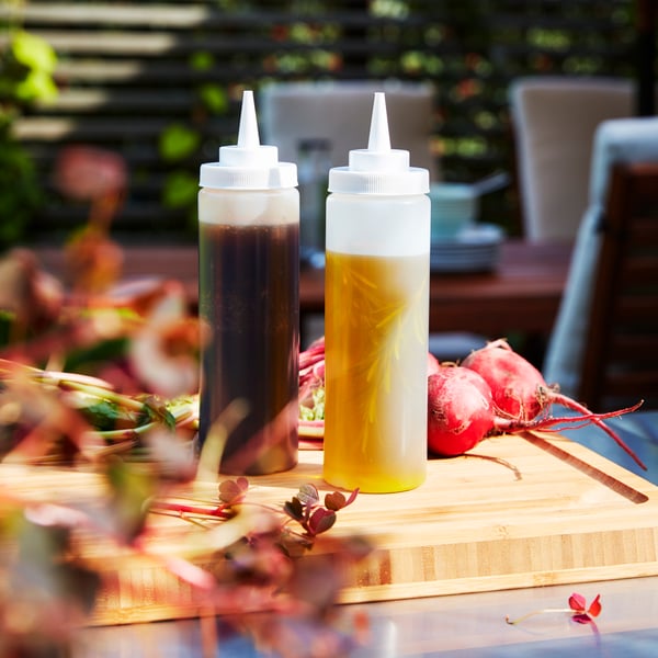 Deux flacons souples GRILLTIDER en plastique transparent contenant des condiments et posés sur une planche à découper en bois à côté d’aliments.