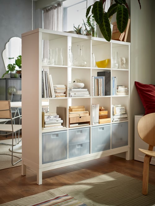A living room with external lighting featuring a white KALLAX shelving unit containing books, glassware, and storage boxes.