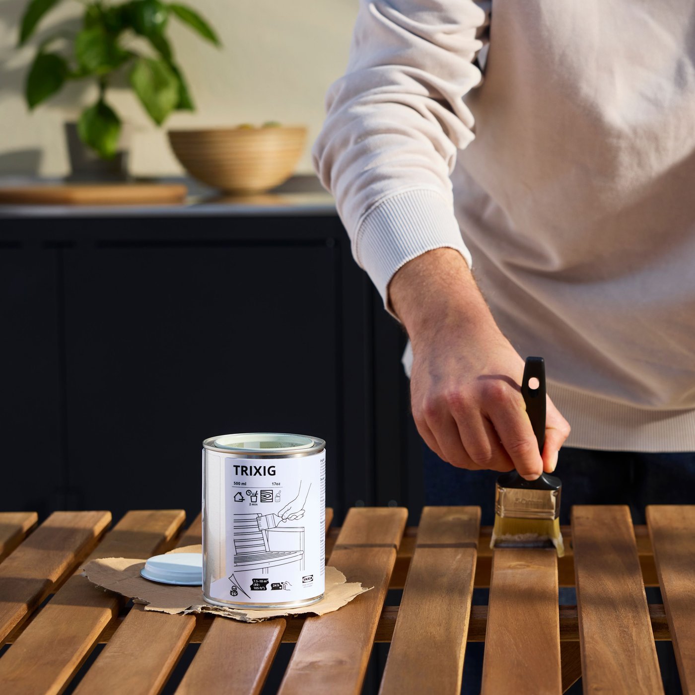 A person is re-staining a wooden outdoor table with a brush next to a can of TRIXIG outdoor wood stain.
