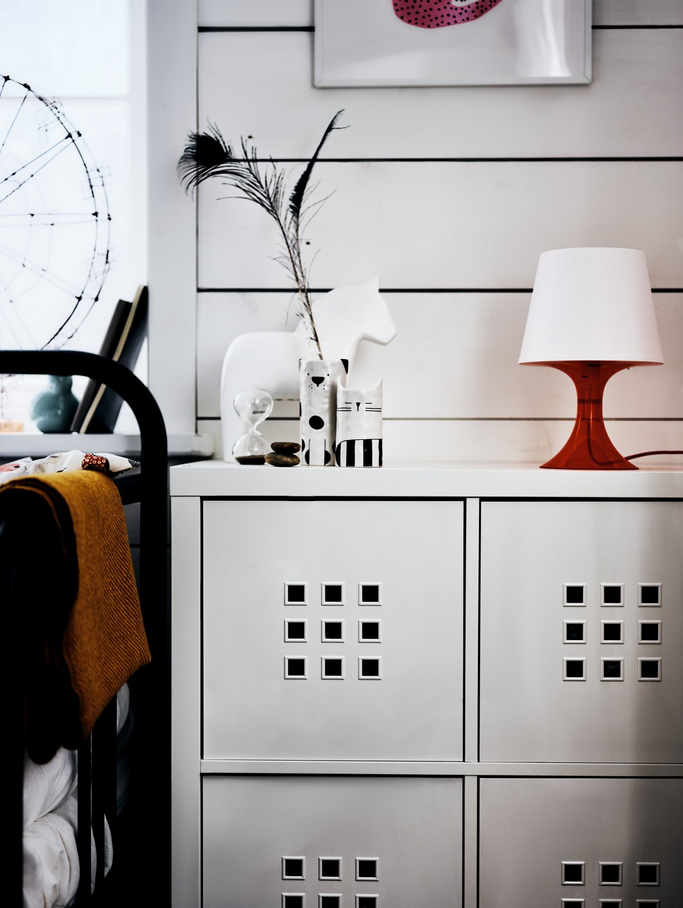 A KALLAX shelving unit with a red lamp, a white model horse and a vase on it, beside a bed frame, in front of a white wall.