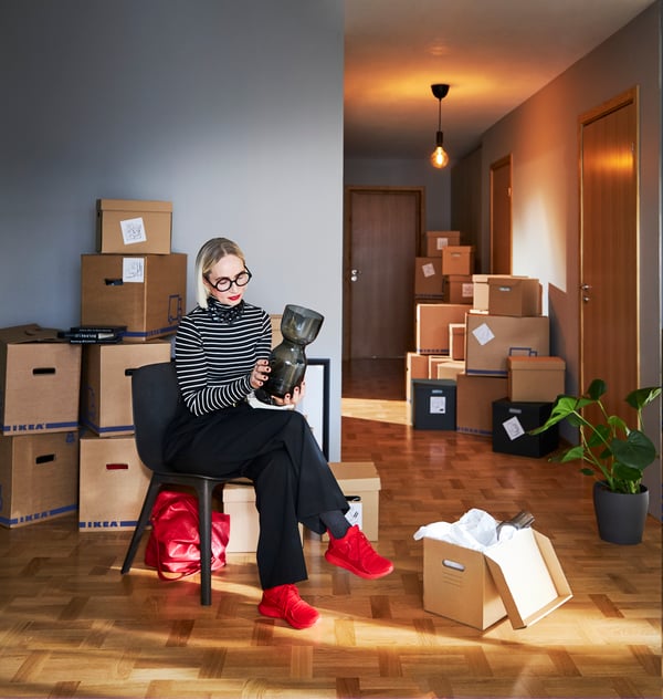 Eine Frau sitzt in einem leeren Raum mit vielen Umzugsboxen. Sie hat rote Schuhe an und hält eine Vase in der Hand.