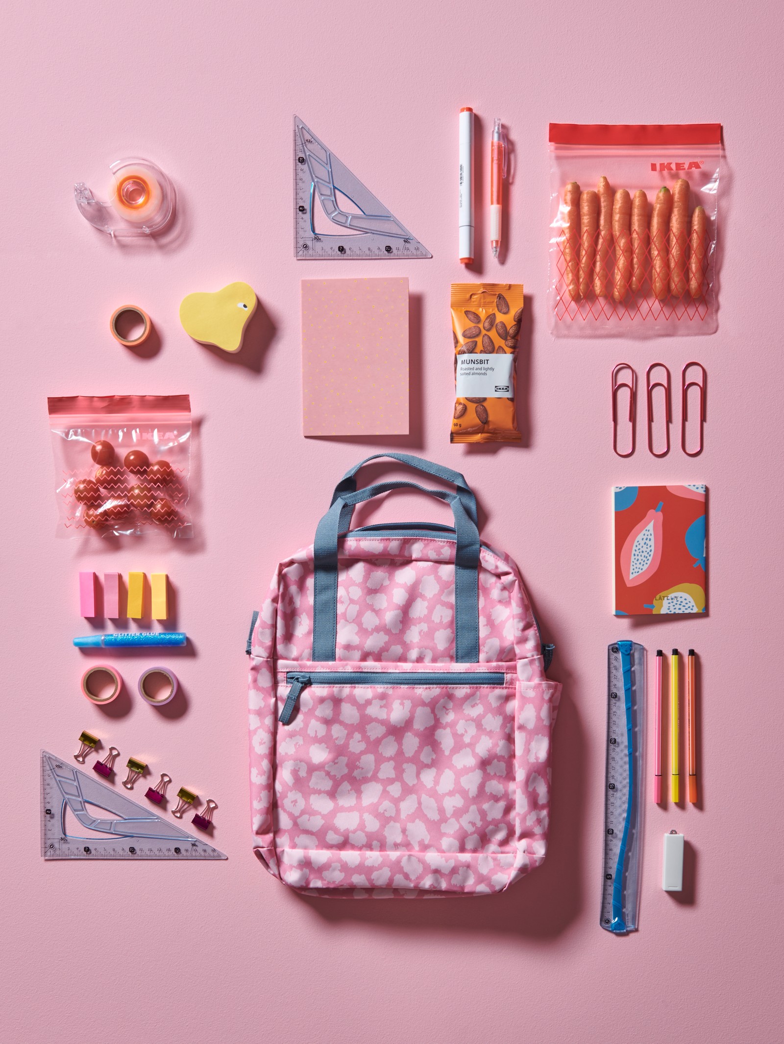 На розовом полу лежит рюкзак с рисунком в розовой гамме, вокруг разложены школьные принадлежности: ручки и скрепки.