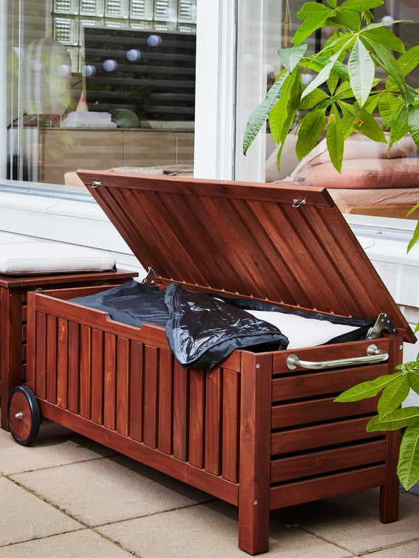 Two benches in acacia wood in front of windows. One bench lid is open and a storage bag filled with cushions is inside.