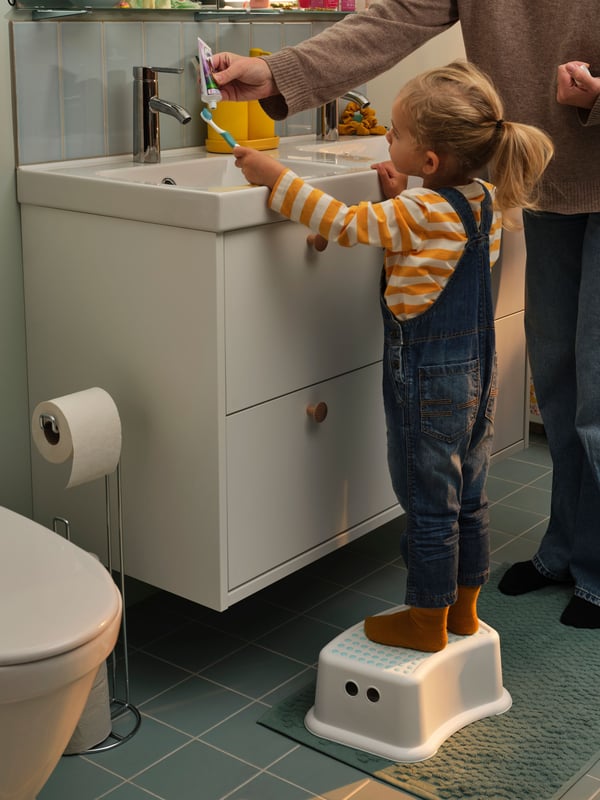 A parent helps a toddler brushing the teeth, the toddler standing on FÖRSIKTIG children’s stool to reach up to the sink.