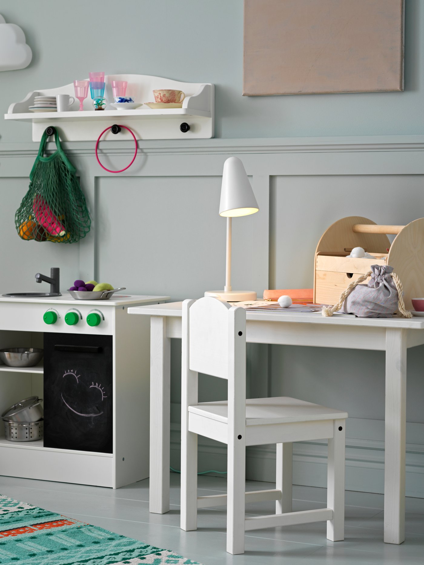 Una silla y una mesa para niños con una lámpara encima, junto a una cocina de juguete con un estante de pared y una lámpara de pared encima.