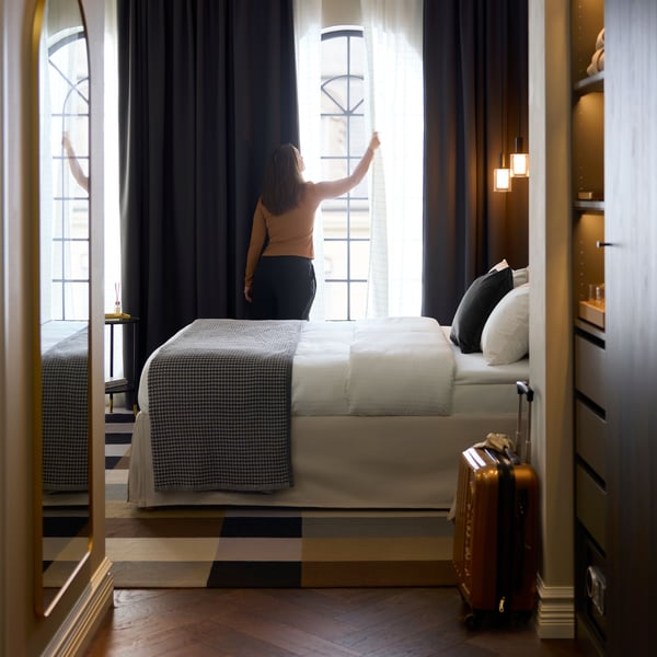 A person is by dark grey KORGMOTT block-out curtains that partially cover sheer curtains on a large, sunny hotel room window.