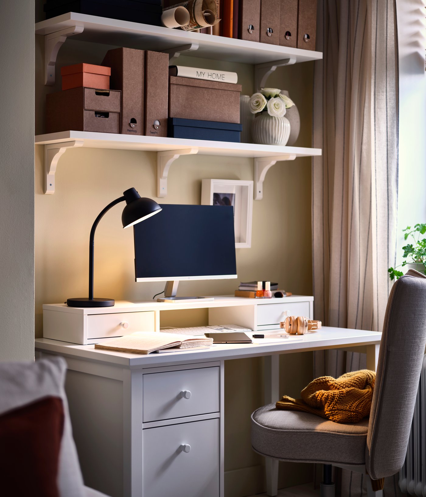 A corner with a study space centred around a white GULLABERG desk with a drop-file drawer, softly lit by a desk lamp.