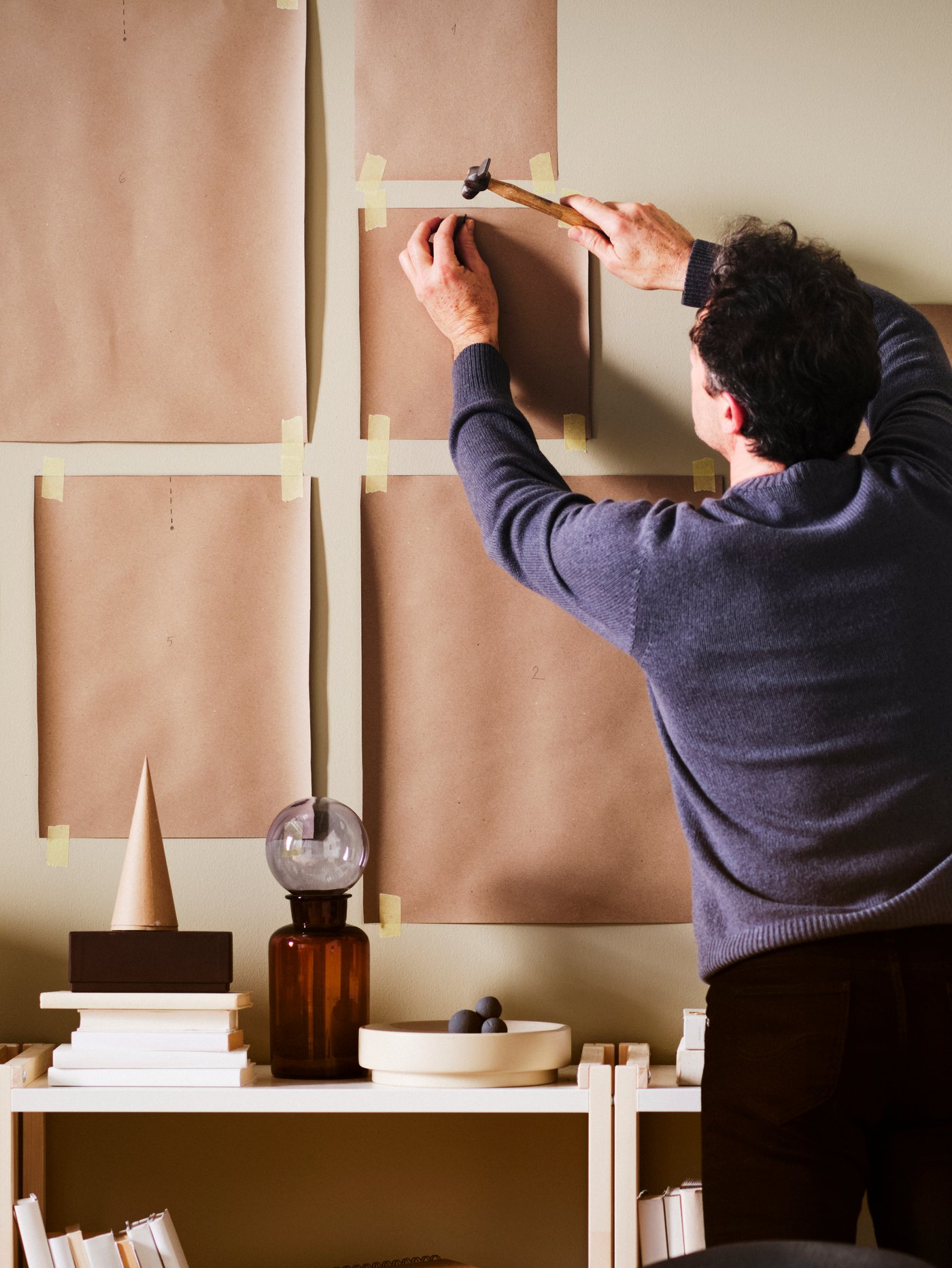 A man driving nails through brown paper templates taped to a wall above a row of white/aspen EKENABBEN open shelving units.