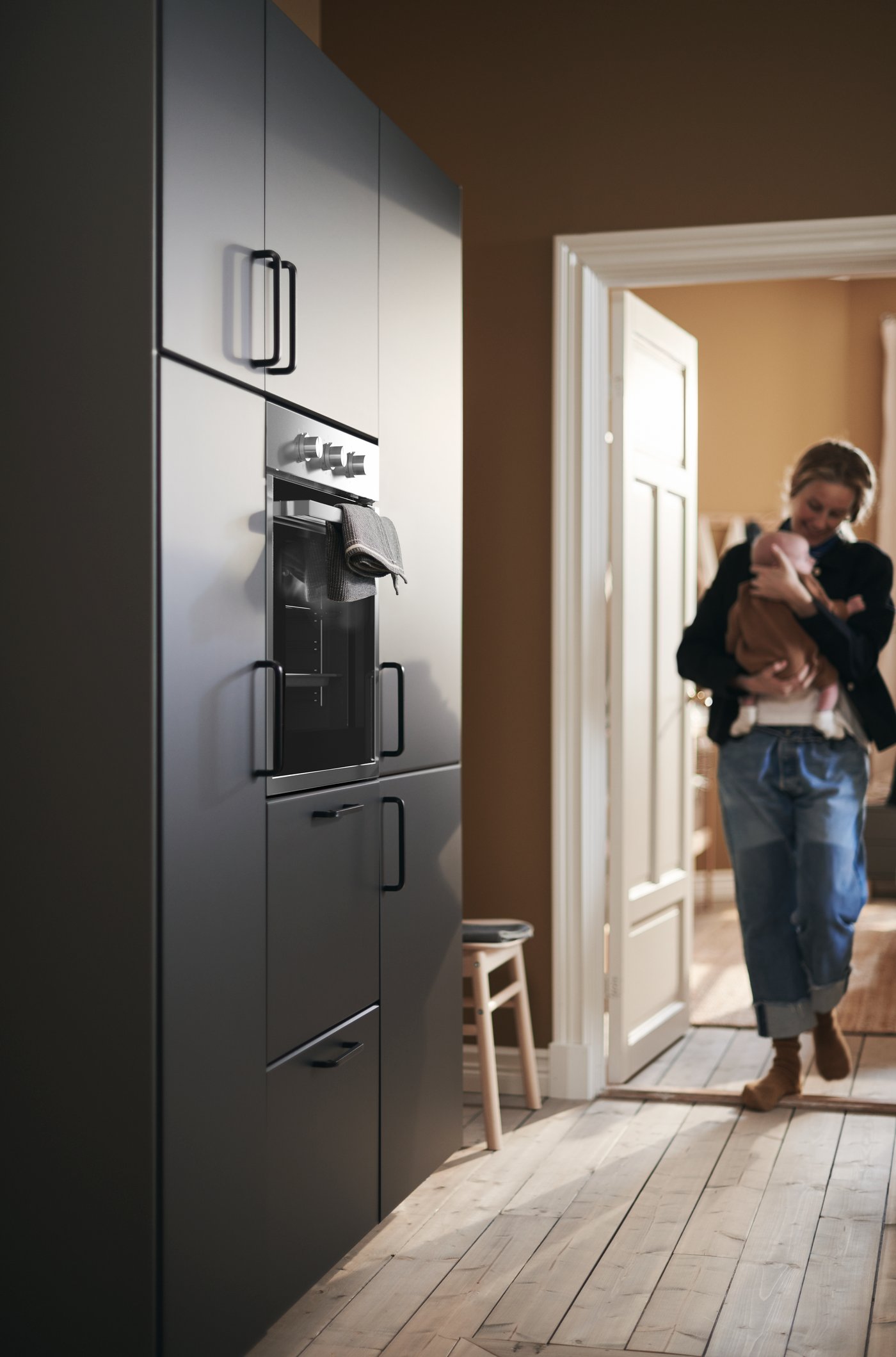 أم تحتضن طفلها وتدخل المطبخ وتقترب من حائط به خزانات مع واجهات KUNGSBACKA.