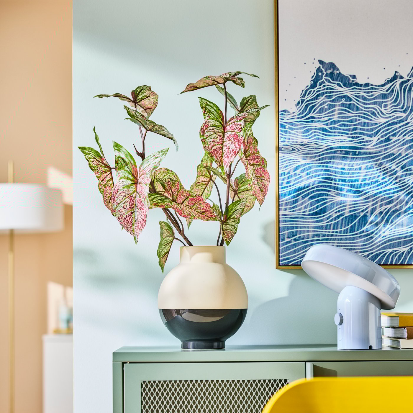 Een zwart-witte GLANSGRAN vaas met roze/groene kunstbladeren erin op een groene kast voor een blauwe muur in de woonkamer.