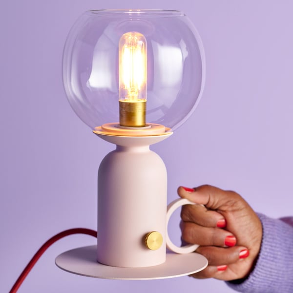 En dimbar ljusrosa/röd ÅSKMULLER bordslampa hålls upp mot en ljuslila bakgrund.