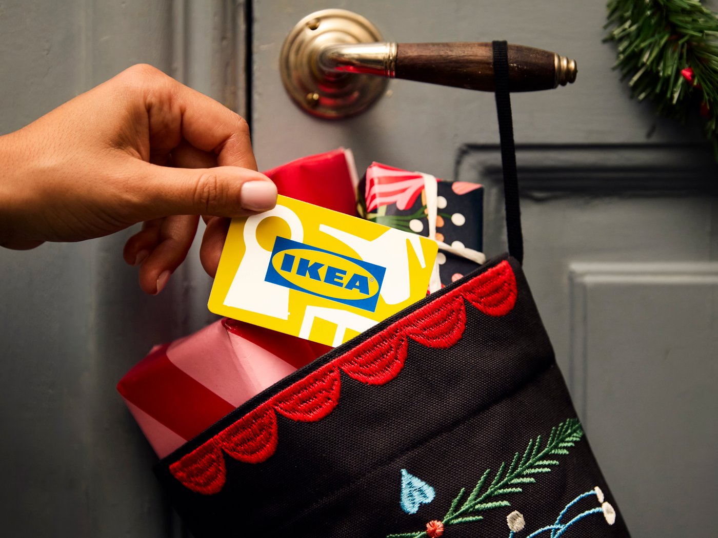 A hand places an IKEA gift card into a festive black stocking with red trim hanging on a doorknob, alongside wrapped gifts, conveying holiday cheer.