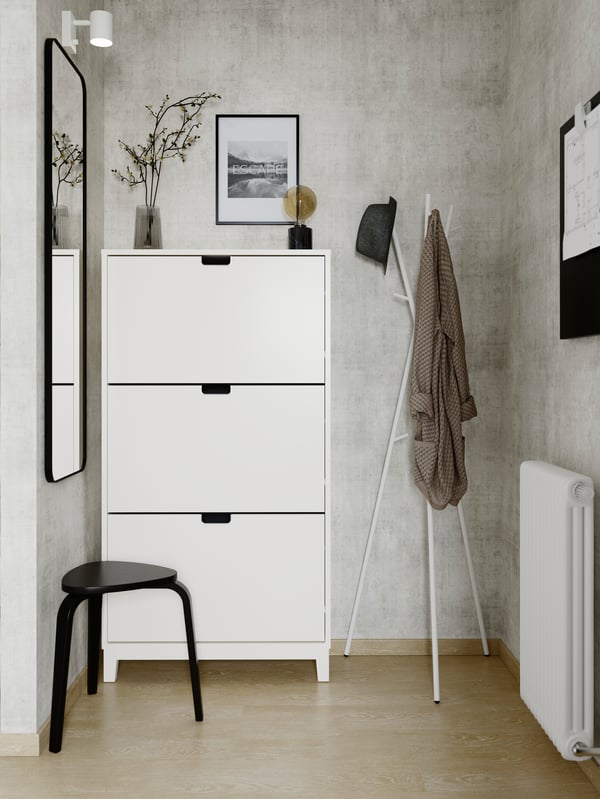 The grey-walled corner of an entryway has a white STÄLL shoe cabinet with 3 compartments by a mirror, stool and coat stand.