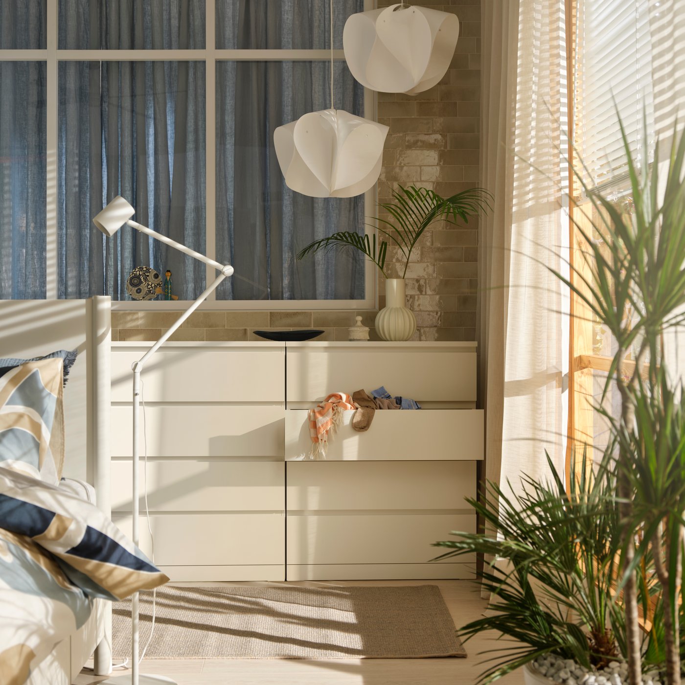 Ett solbelyst och modernt sovrum med en vit byrå, en golvlampa och en stor krukväxt som skapar en lugn och inbjudande atmosfär.