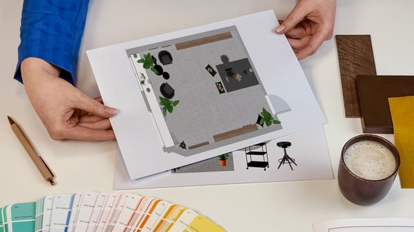 Projektantka wnętrz IKEA prezentująca kartki z wydrukiem rzutu z góry zaprojektowanego pomieszczenia biznesowego. Obok na blacie leży wzornik kolorów i próbki materiałów.