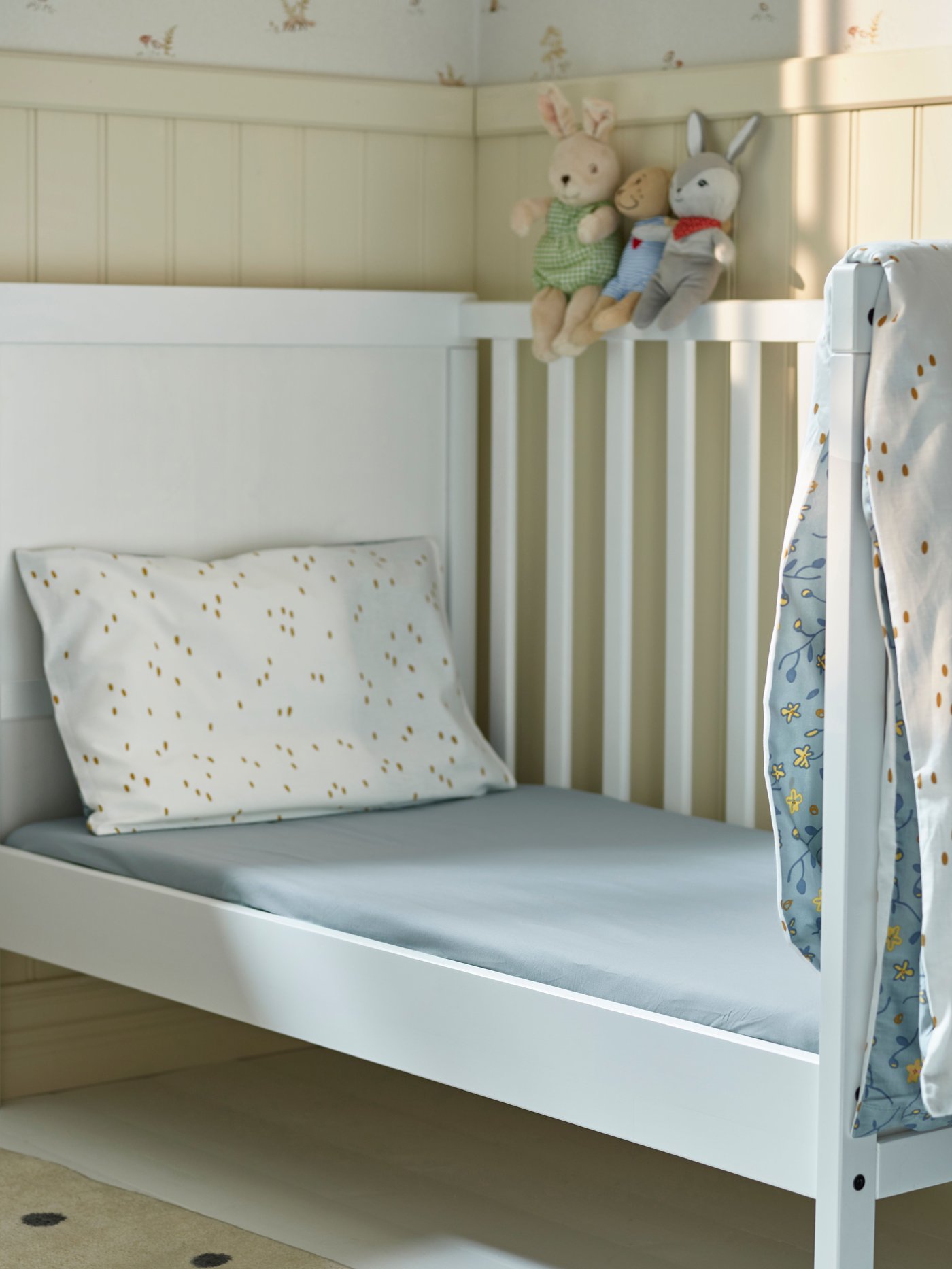 White cot with blue sheets and white pillowcases with small patterns. Three soft toys sit on the bed's backrest and a blanket hangs over the side.
