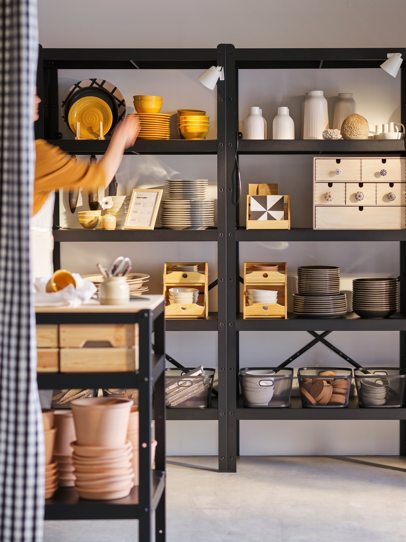 A shop keeper standing to the left of a set of high BROR shelves adjusts items on display on the top shelf.