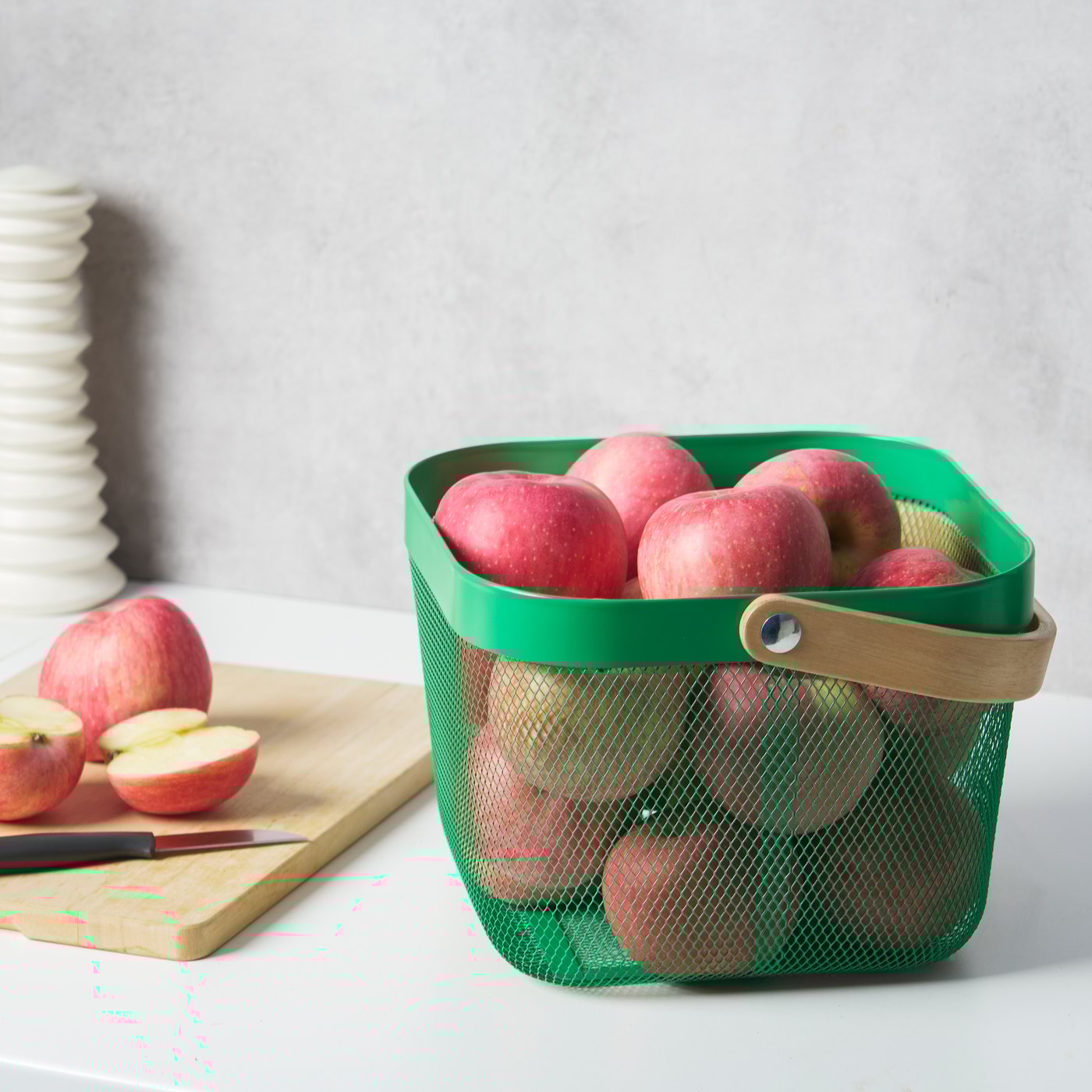 Tasolla on leikkuulaudalla kaksi omenaa sekä vihreä RISATORP-kori täynnä punaisia omenoita.