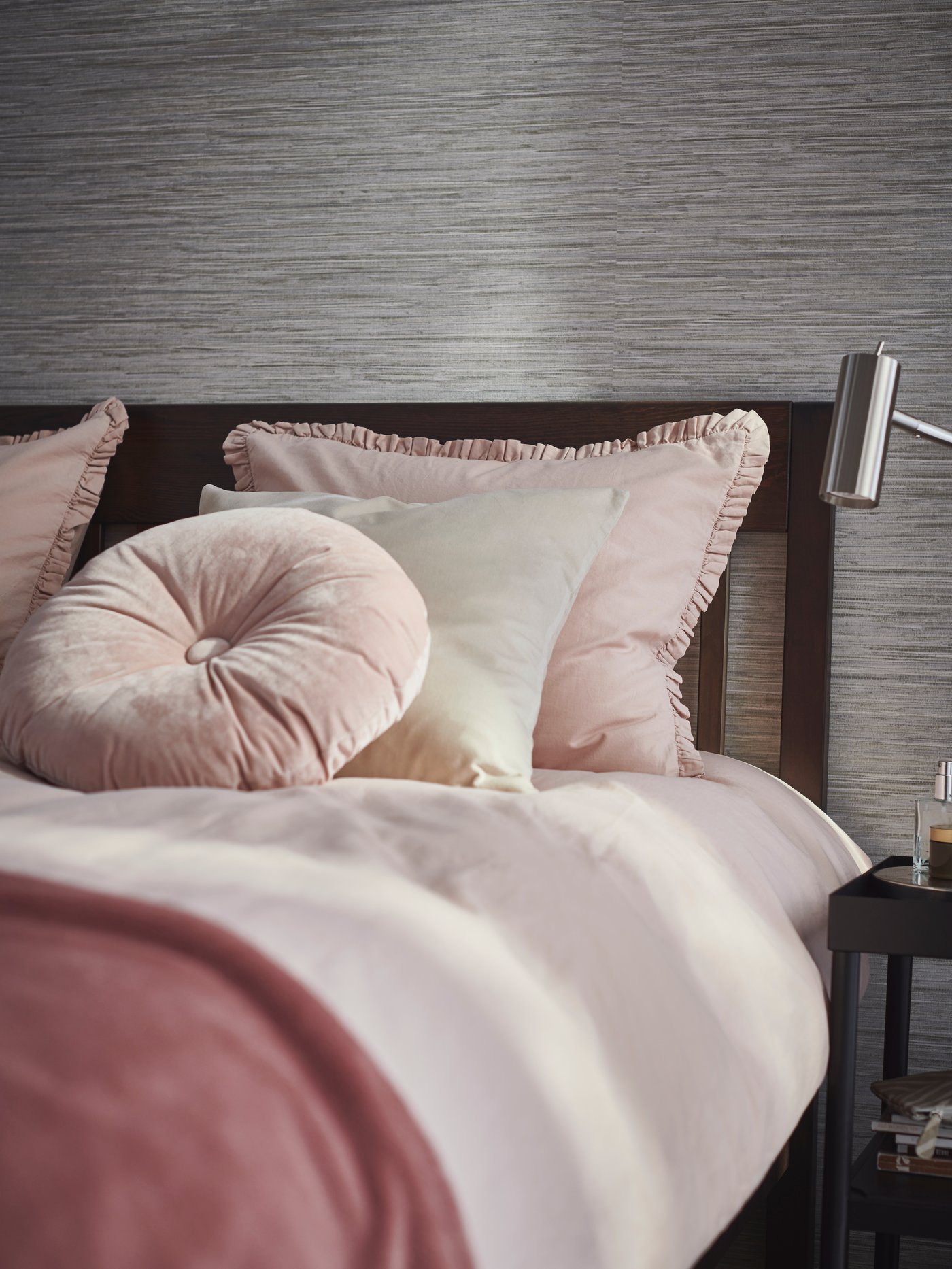 في غرفة نوم، توجد وسادة KRANSBORRE وغطاء وسادة SANELA وطقم غطاء لحاف KRANSKRAGE وردي على سرير مرتب.