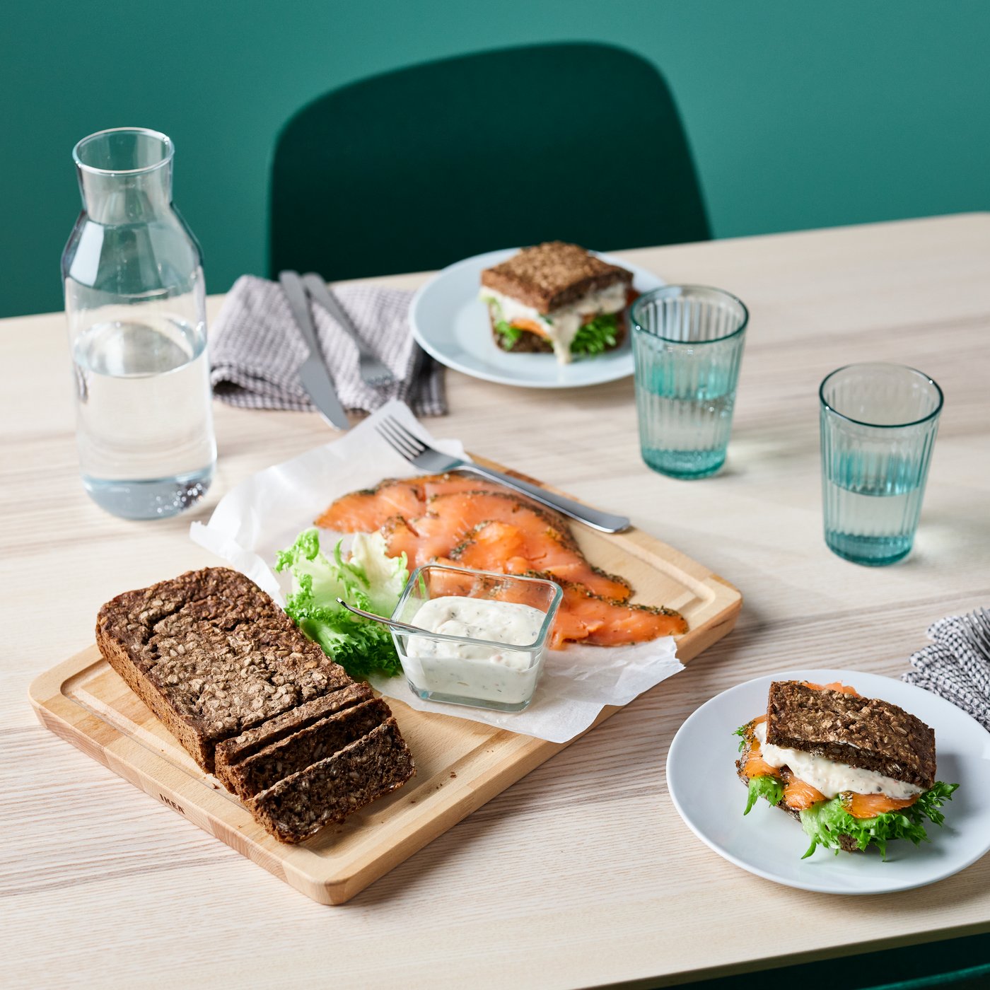 Gedeckter Tisch mit Sandwich-Tellern (dunkles Brot, Salat, Räucherlachs), einem Holzbrett mit Brotscheiben, Räucherlachs und cremiger Sauce, leeren Gläsern, einer Wasserkaraffe und gefalteten Stoffservietten vor einer grünen Wand und mit grünem Stuhl im Hintergrund.