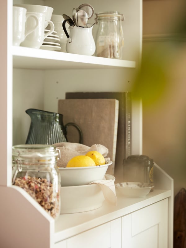 Two shelves of a white HAUGA high cabinet with 2 doors holding tableware, books and food including two lemons.