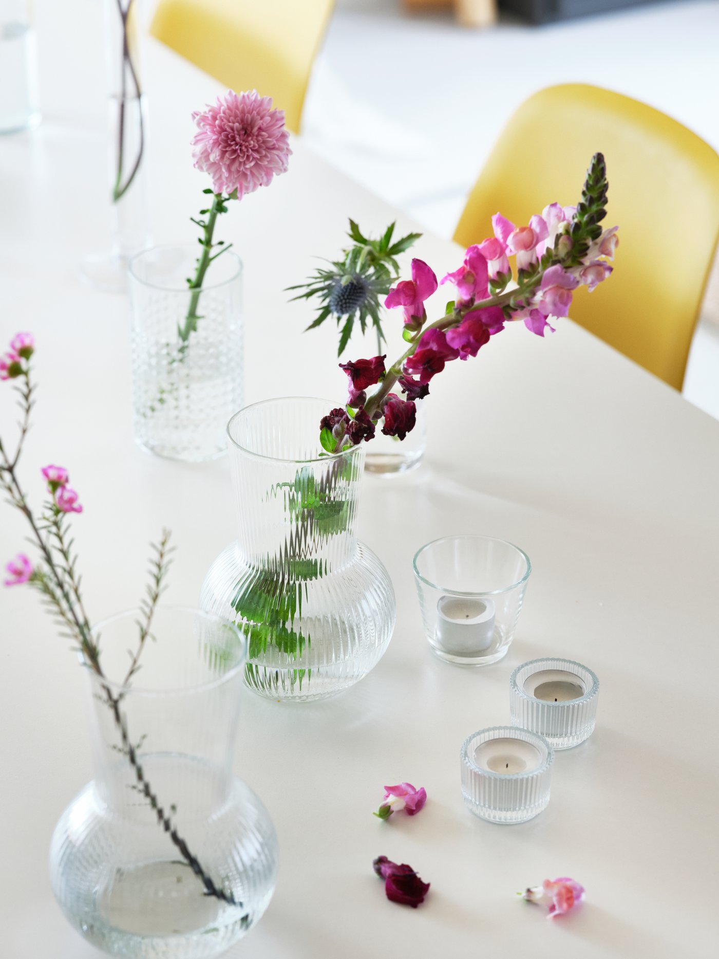 Varios floreros con tallos de flores y portavelas sobre una mesa blanca. Hay sillas amarillas junto a la mesa.