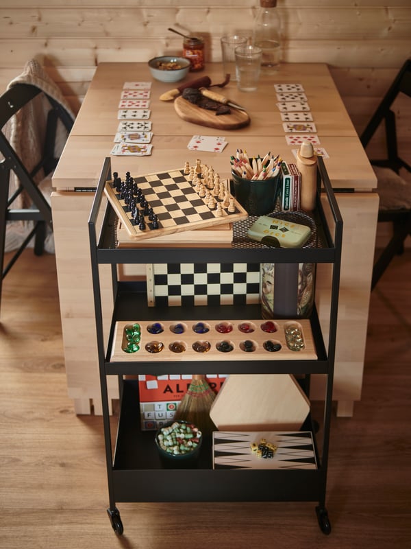 NORDEN klaffbord uppdukat för sällskapsspel med bland annat schack, och hyllor under bordet fyllda med spelpjäser och tillbehör.