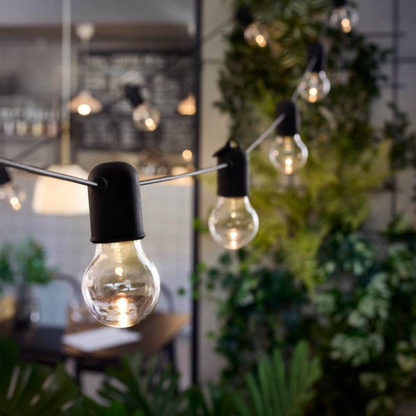 An outdoor area with greenery in the background shows a black SVARTRÅ LED lighting chain with 20 bulbs strung above seating.