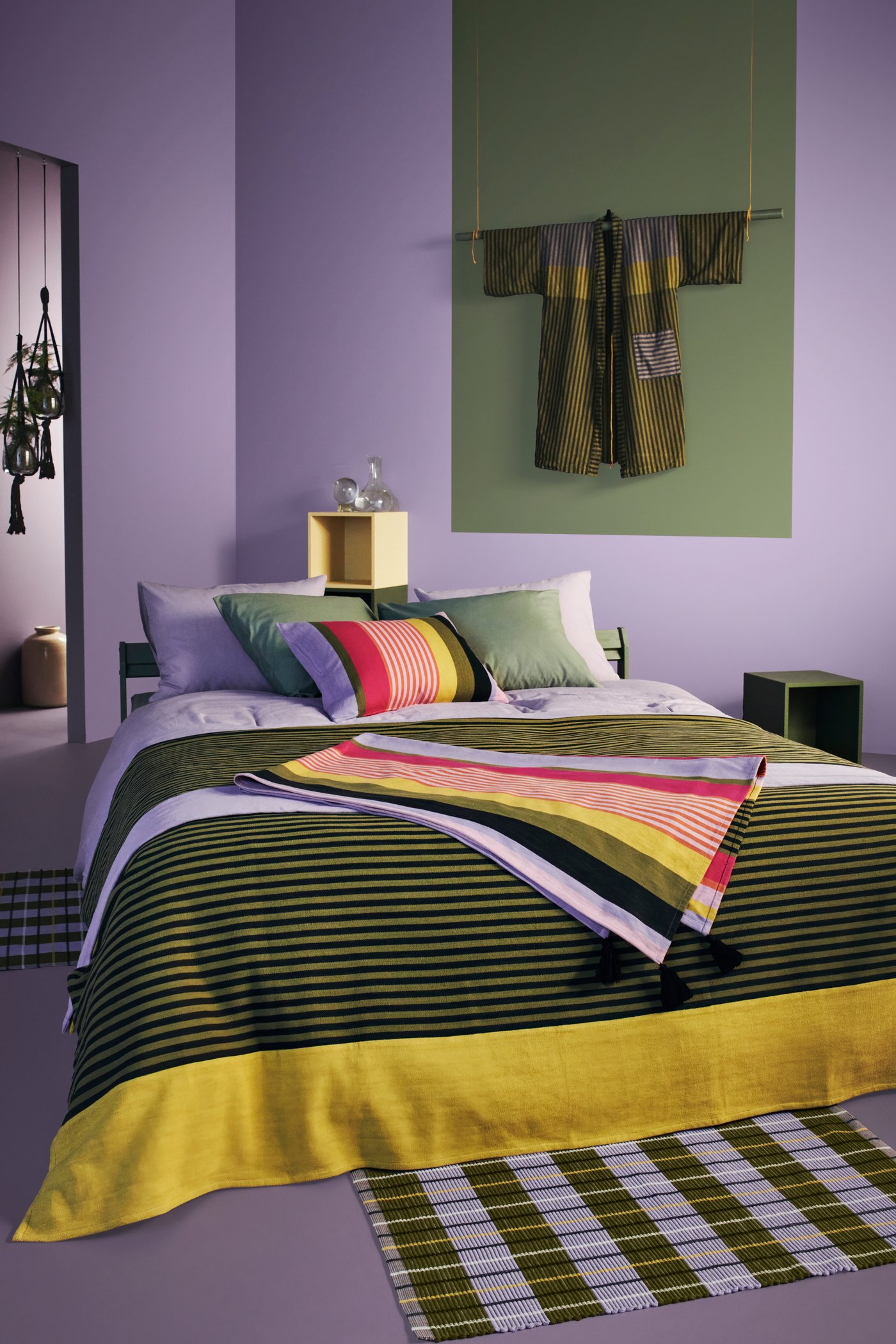 غرفة باللون الأرجواني وسرير مغطى بمفرش MÄVINN مخطط بألوان متعددة. روب حمام MÄVINN معلق على الجدار.